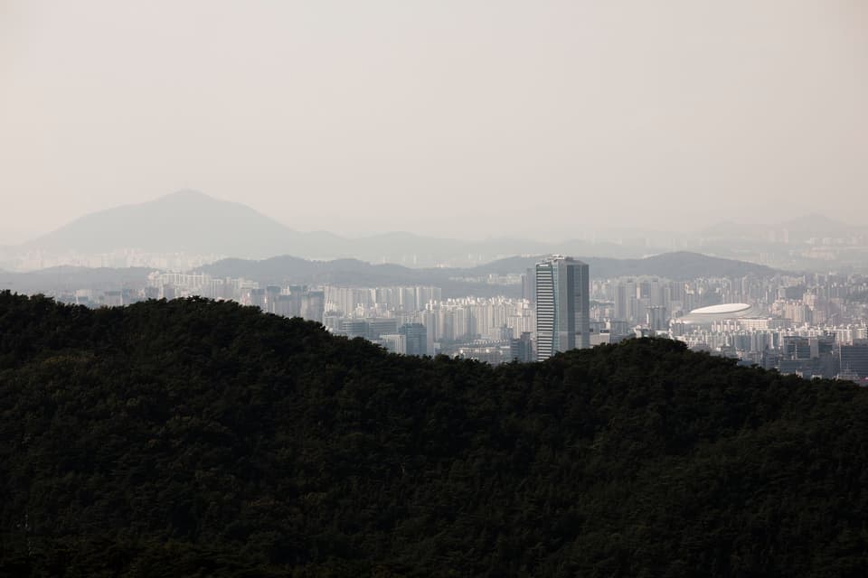 Aerial skyline of Seoul from forested hillsides, featuring distant mountains.