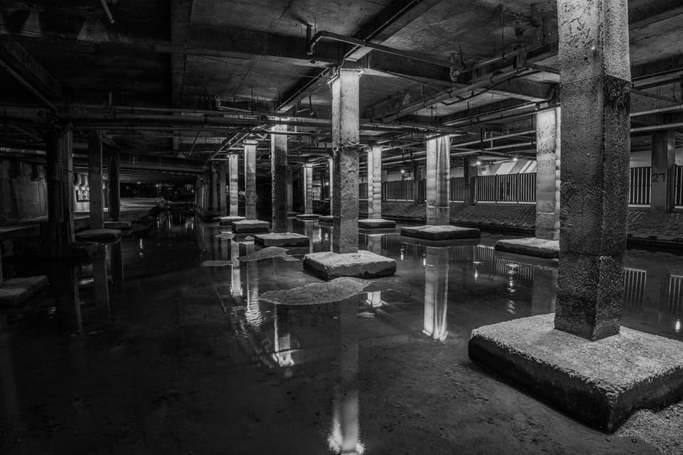 Atmospheric black and white photo of an abandoned underground space with concrete columns and water reflections.