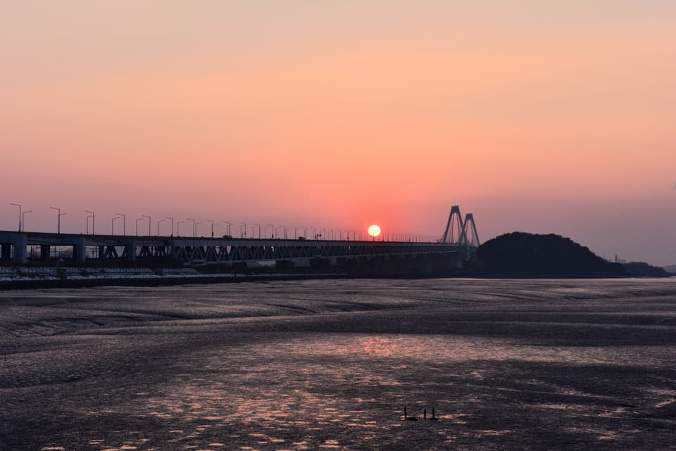 Captivating view of Yeongjong Bridge during sunset in Incheon, South Korea with serene waters and a warm orange sky.