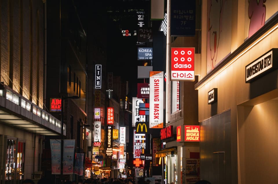 Dynamic night view of Seoul's bustling shopping district featuring various store signs and urban lights.