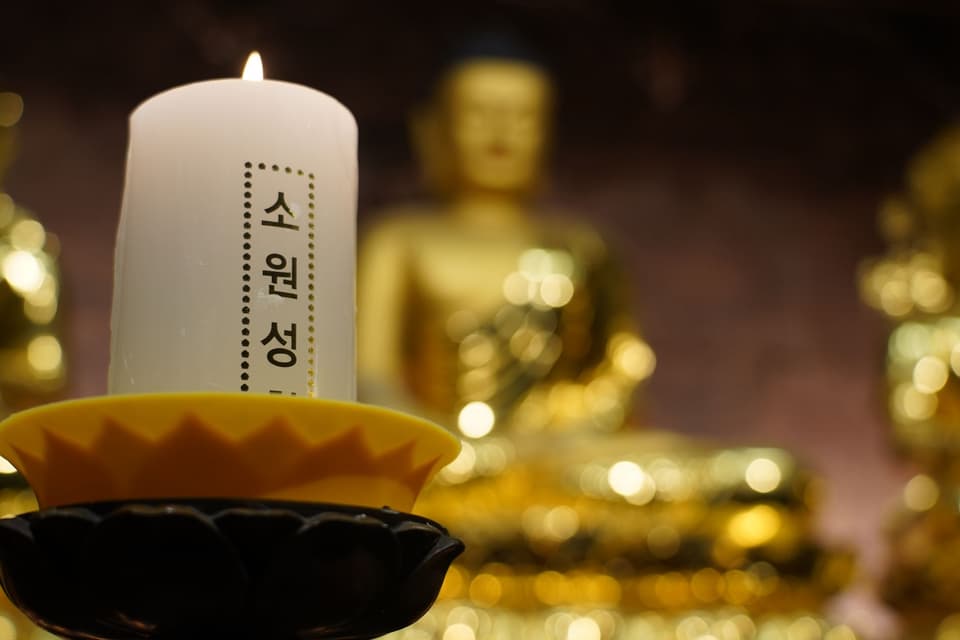 A lit candle with Korean text in front of a golden Buddha statue, Seoul temple.