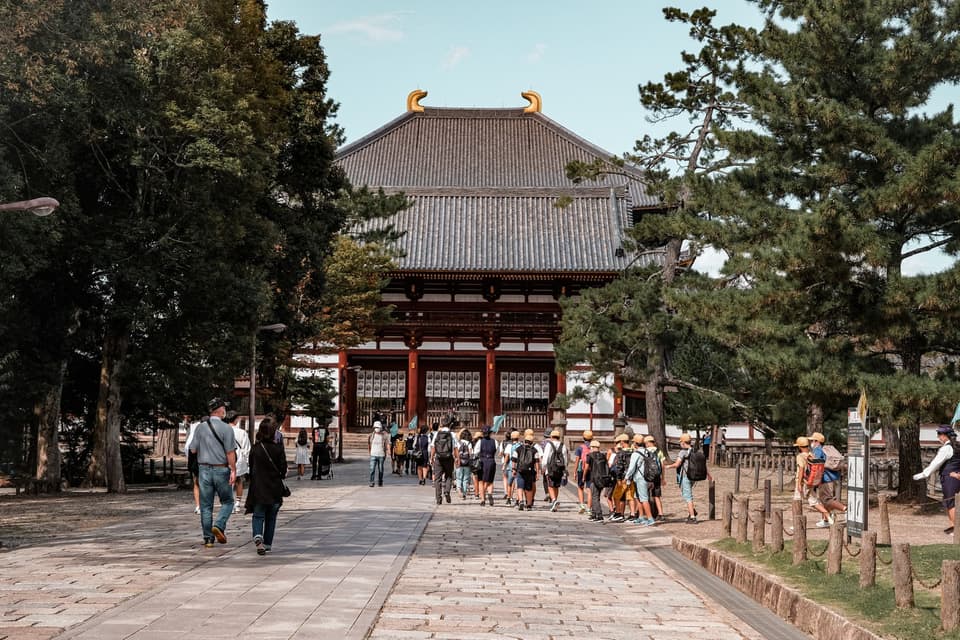 Tourists walking towards Todai-ji Temple in Nara, Japan, surrounded by trees and historical architecture.