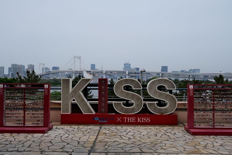 KISS sign and love padlocks at Aqua City Odaiba with Tokyo's skyline and Rainbow Bridge in the background.