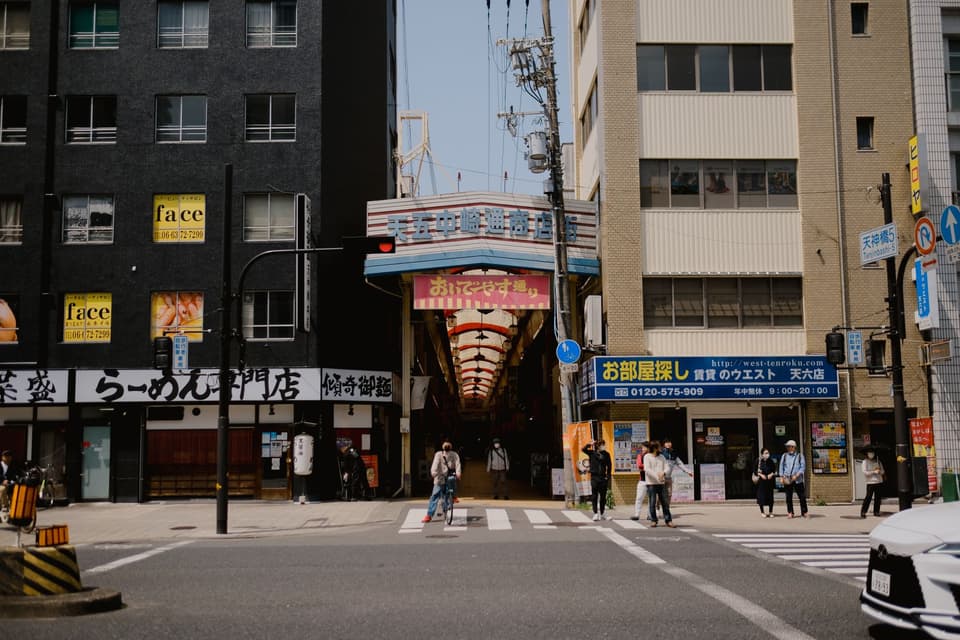 Street view of bustling Osaka with people crossing, shops, and arcade entrance.