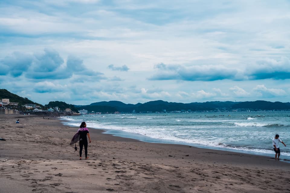 People strolling along Kamakura beach, Japan on a cloudy day, with scenic ocean waves and distant hills.